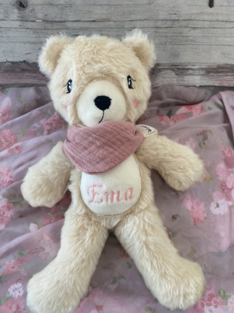 Personalisierter Kuschelbär aus weichem Langhaarplüsch mit rosa Musselin Tuch und dem Namen EMA auf dem Bauch, liegend auf einer rosa Blumendecke