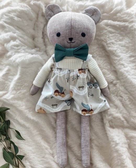 Handgemachte Stoffpuppe in Bärenform aus Leinen,40 cm gross, mit grüner Fliege, weissem Oberteil und gemusterten Latzhosen liegend auf weisser Decke.