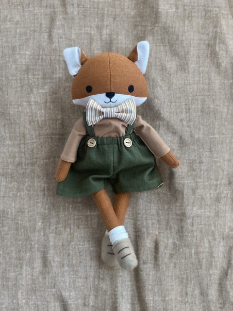 Handgemachtes Stofftier in Form eines Fuchses, gekleidet in einer grüne Latzhose, ein braunes Oberteil und einer gestreifte Fliege, liegend in einer Holzkiste mit Holzwolle auf einem Holzboden.

