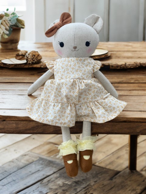 Handgemachte Stoffpuppe in Form eines Bären mit Schleife am Ohr, geblümtem Kleid und kleinen Stoffschuhe, sitzend auf einem Holztisch 