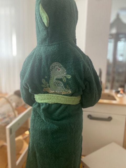 Kleines Kind in einem grünen Dino-Bademantel mit Kapuze und Zacken, von hinten fotografiert. Der Bademantel hat ein Dino Motiv am Rücken.
