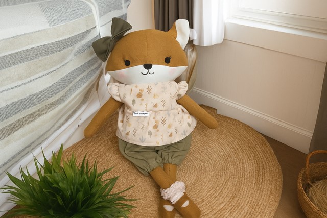 Stofftier in Form eines Fuchses, das auf einem runden Teppich sitzt. Das Kuscheltier trägt eine rosa Bluse mit Waldmotiv, grüne Pumphosen, braune Schuhe mi rosa Socken und eine grüne Schleife am Ohr.

