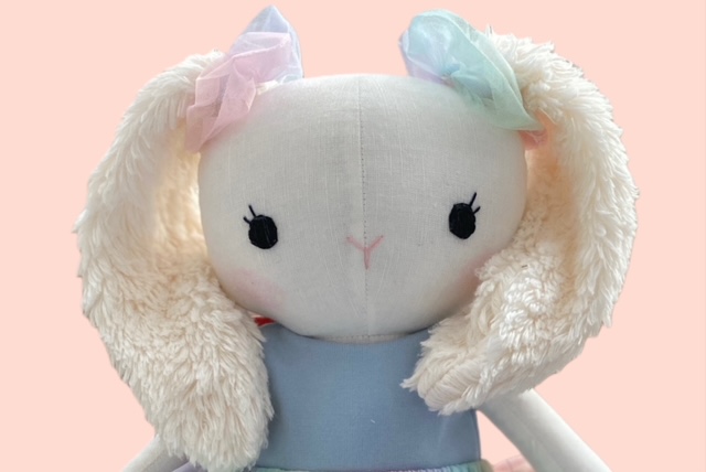 Nahaufnahme einer plüschigen Stoffhasenpuppe mit wichen, langen Ohren, pastellfarbenen Schleife und bestickten Gesicht.  