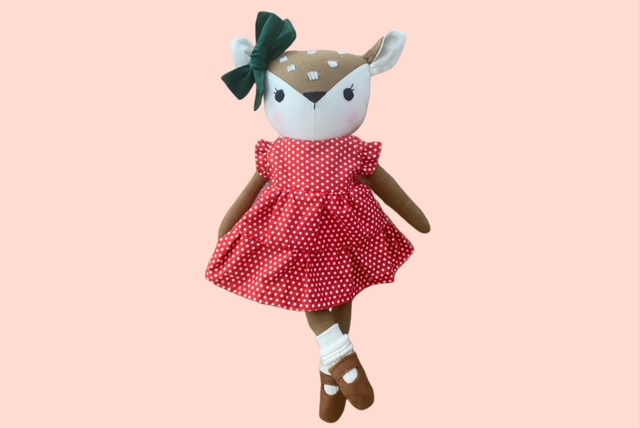 Stoff-Rehpuppe mit dunkelgrüner Schleife und rotem Kleid mit weissen Punkten, die mit überkreuzten Beinen posiert.