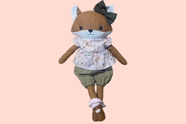 Stoff- Fuchspuppe mit dunkelgrüner Schleife, Waldmotiv- Oberteil und locker sitzenden Hose, die mit gekreuzten Beinen posiert.