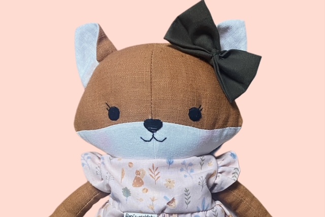 Nahaufnahme einer Stoff-Fuchspuppe mit dunkelgrüner Schleife, gestickten Gesicht und einem Oberteil mit zarten Waldmotiven.