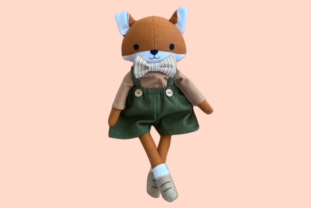 Stoff- Fuchspuppe mit kurzen Latzhosen mit Knöpfen, beigefarbenem Oberteil und gestreifter Fliege, die mit gekreuzten Beinen posiert.