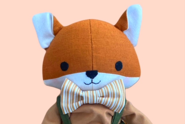 Nahaufnahme einer Stoff- Fuchspuppe mit gestreiften Fliege und einem gestickten Gesicht vor pastellfarbenem Hintergrund.