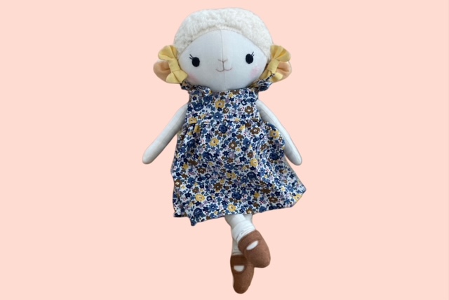 Stoff-Schafpuppe im geblümten Kleid und braunen Schuhen, die mit gekreuzten Beinen posiert.