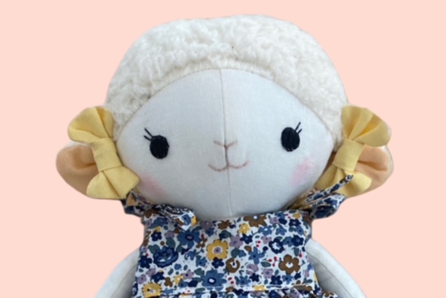 Nahaufnahme einer Stoff-Schafpuppe mit flauschigem Kopf, geben Schleife an den Ohren und bestickten Gesicht, die ein geblümtes Kleid trägt.