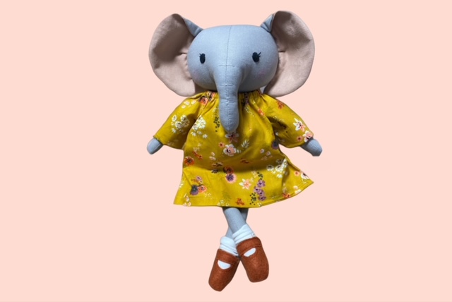 Stoff-Elefantenpuppe in einem gelben, geblümten Kleid und braunen Schuhen, die mit gekreuzten Beinen posiert.