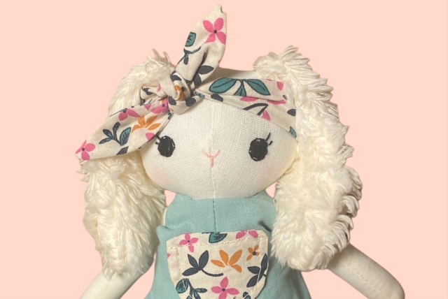Nahaufnahme einer Stoff-Hasenpuppe mit Flauschiegen Ohren, floralem Haarband und mintfarbener Latzhose mit Blumen-Tasche 