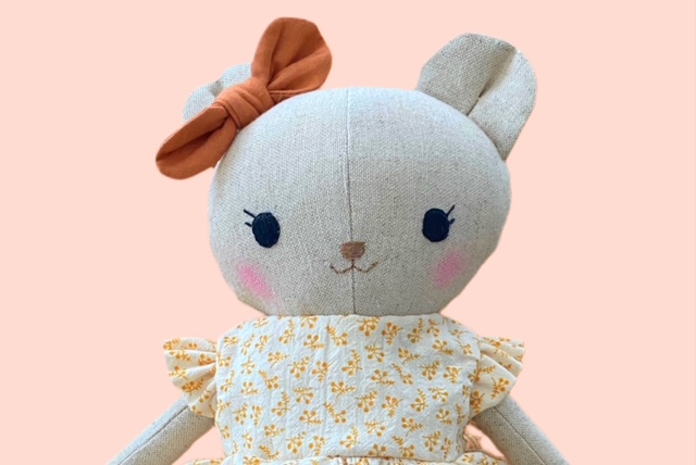 Nahaufnahme einer Stoff-Bärenpuppe mit brauner Schleife und gelbem Blumenkleid, das Gesicht ist gestickt.