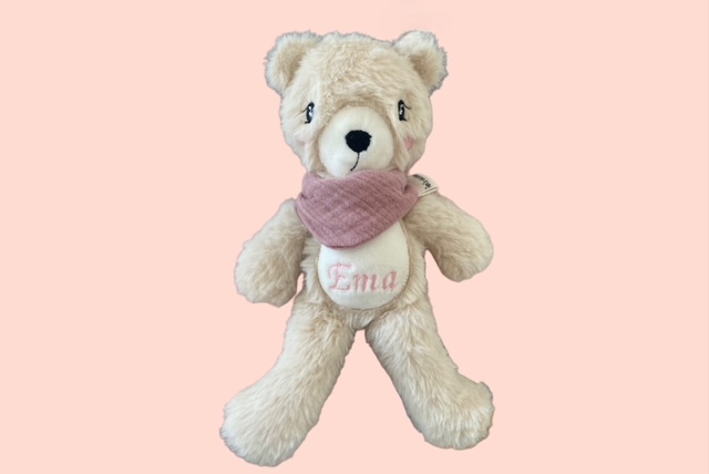 Cremefarbener Stoffbär mit rosafarbenem Schal und besticktem Namen auch dem Bauch, auf dem Name Ema steht.