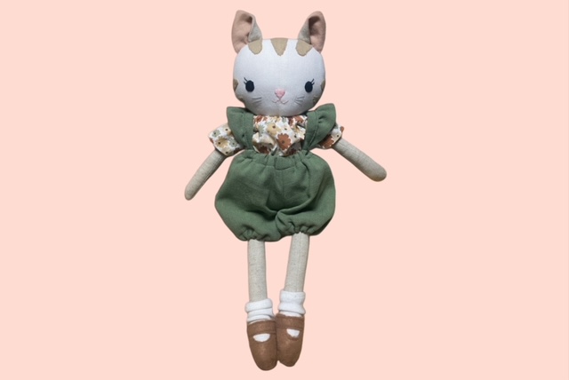 Stoff-Katzenpuppe mit Blumenoberteil, grüner Pumphosen und braunen Schuhen vor pastellfarbenem Hintergrund.