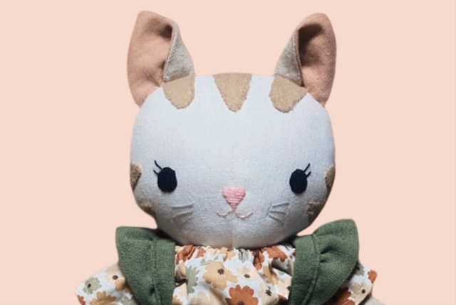 Nahaufnahme einer Stoff-Katzpuppen mit aufgesticktem Gesicht, beigen und braunen Flecken und einem Oberteil mit Blumenmuster.