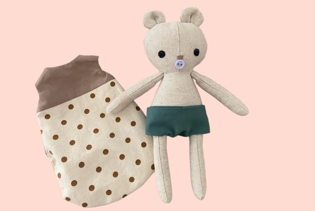 Kleiner Stoff-Bärenpuppe mit grüner Stoffwindel neben einem gepunkteten Schlafsack aus Stoff