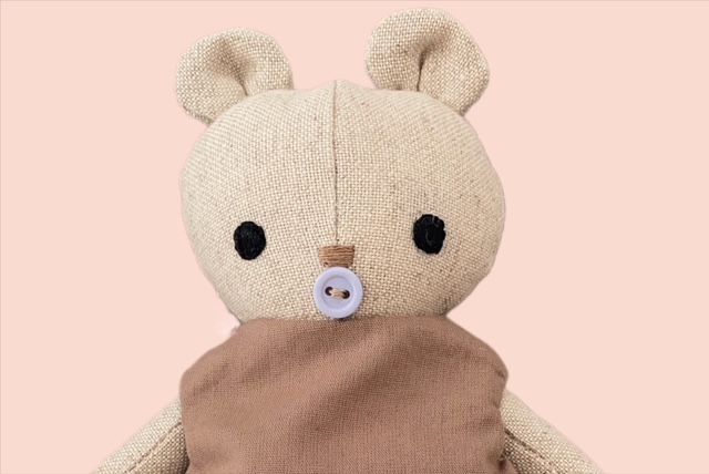 Nahaufnahme einer kleiner Stoff-Bärenpuppe mit gesticktem Gesicht und einem Knopfschnuller, die ein einfaches braunes Oberteil trägt.