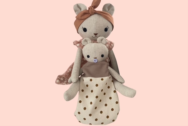 Stoff-Bärenmama in rosafarbenem Blumenkleid mit Haarband, die ein kleines Bärenbaby in gepunkteten Schlafsack vor sich trägt.