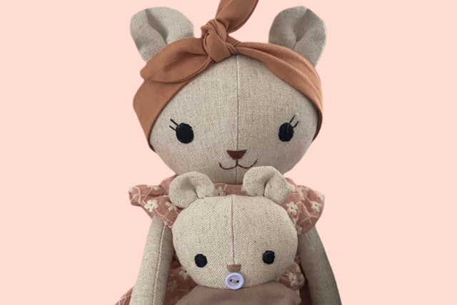 Nahaufnahme einer Stoff-Bärenmama mit braunfarbenem Haarband und rosa Blumenkleid, die ein kleines Stoff- Bärenbaby im Arm hält.