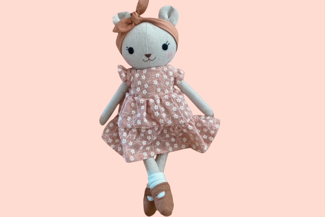 Stoff-Bärenpuppe in rosafarbenem Kleid mit weissen Blüten und passendem Haarband sitzend mit überkreuzten Beinen vor hellem Hindergrund.