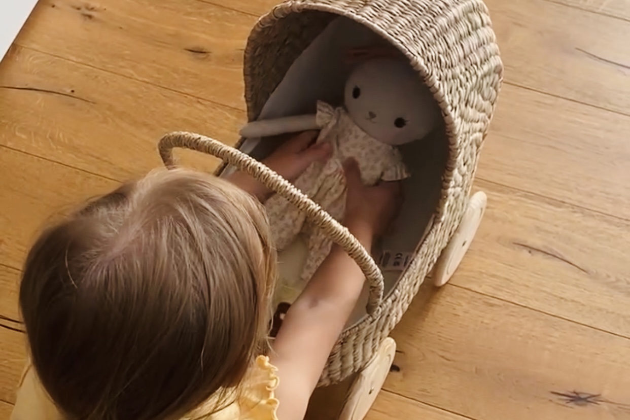 Kleines Kind legt eine handgenähte Stoffpuppe in eine geflochtenen Puppenwagen aus Rattan.