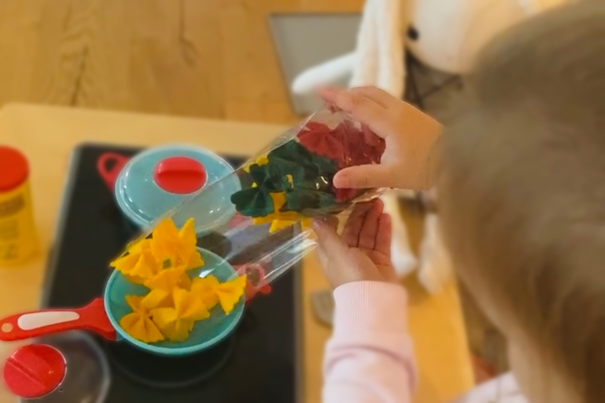 Kind Schüttet bunte Filz-Nudeln in einen kleinen Spielkochtopf, während eine Plüschpuppe daneben sitzt.