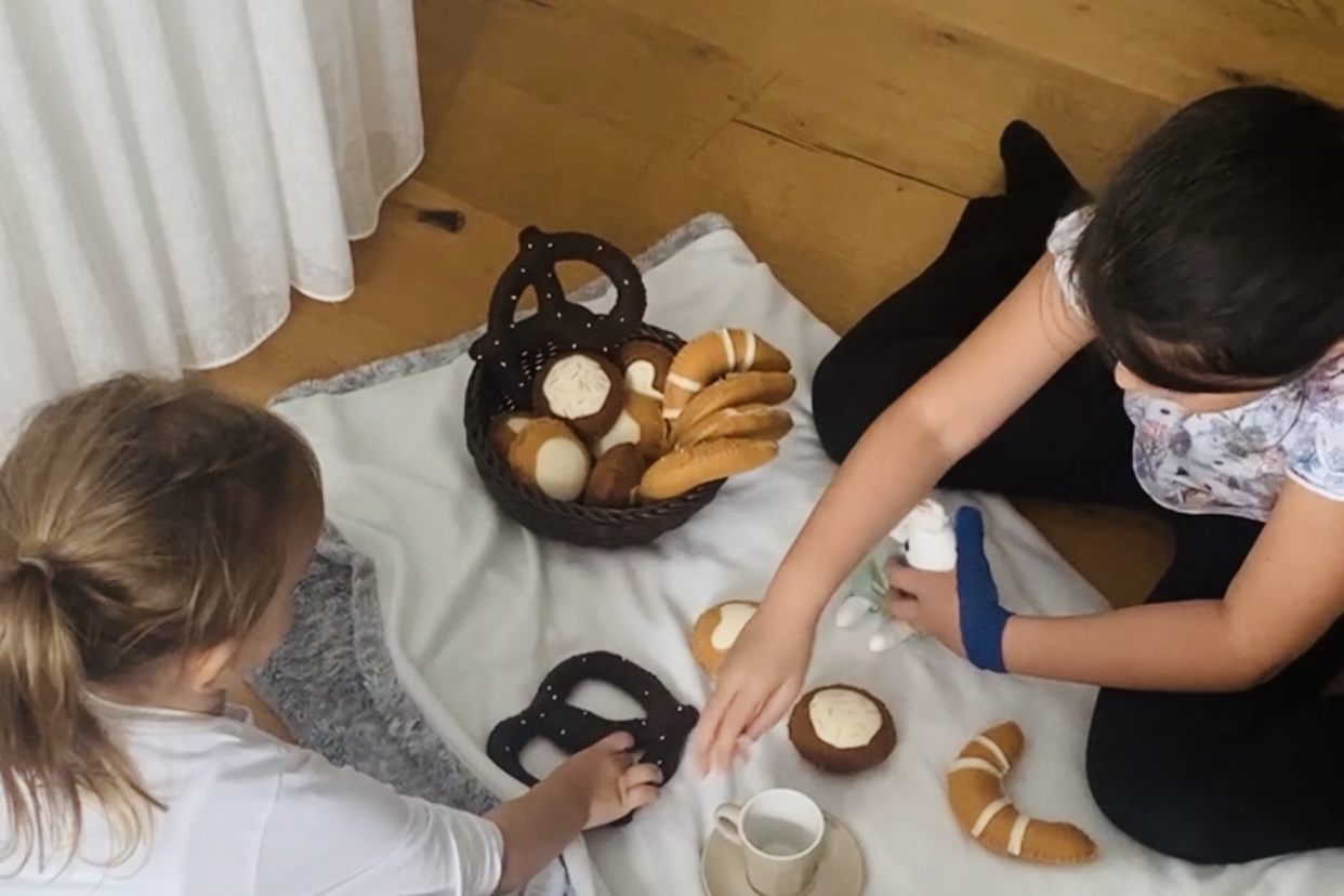 Zwei Kinder spielen gemeinsam Picknick auf einer Decke. In der Mitte steht ein Korb voller Filzbrötchen und Gebäck, während eines der Kinder eine Filzbrezel greift.