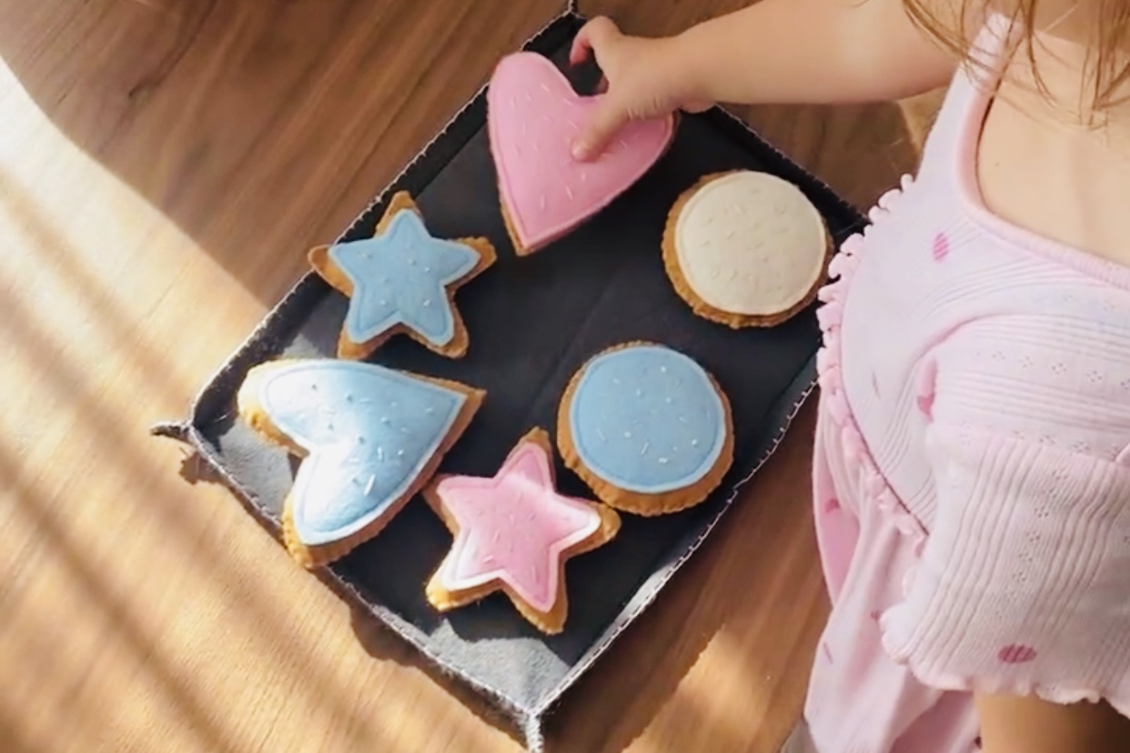 Ein Kind legt bunte Filzkekse in verschiedenen Formen- Herz, Stern, und Kreis auf ein Tablett. Sonnenlicht fällt warm über den Holztisch  