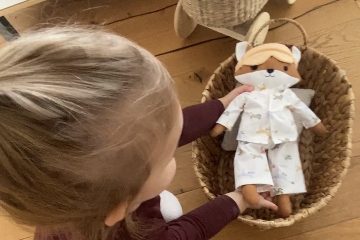 Ein Kind legt eine Stoffpuppe im Pyjama vorsichtig in einen geflochtenen Korb. Die Szene zeigt liebevolles Spielen auf einem Holzboden.