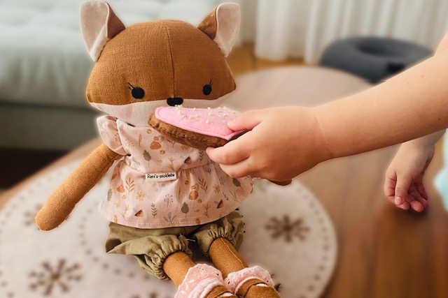 Ein Kind hält einer handgemachten Stoffpuppe in Fuchsform ein rosa Filz-Keks- Spielzeug hin. Die Puppe sitzt auf einem Teppich und trägt ein liebevoll genähtes Ouitfit.