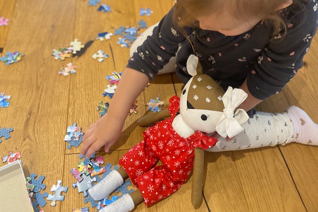 Ein Kind sitzt auf dem Boden und spielt mit einer handgemachten Stoffpuppe mit Weihnachtsoutfit. Rundherum liegen bunte Puzzleteil.