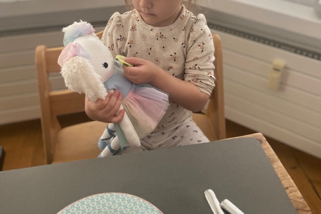 Ein Kind sitzt am Tisch und hält eine handgemachte Stoffpuppe im Ballerina-Outfit in den Armen.