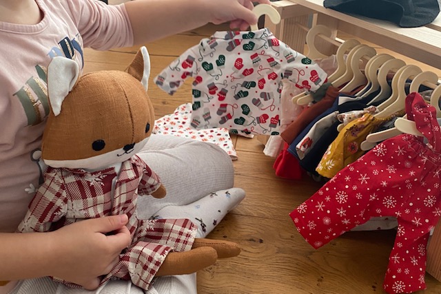 Kind spielt mit einer Stoffpuppe und hält eine Puppenkleid mit Weihnachtsmotiv, daneben eine Kleiderstange mit Puppenkleidung. 