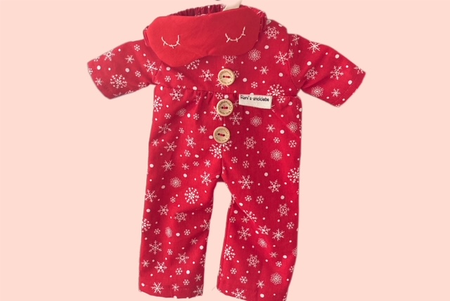 Roter Puppen-Strampler mit weissen Schneeflocken, Knöpfen und passender Schlafmaske auf rosa Hintergrund.