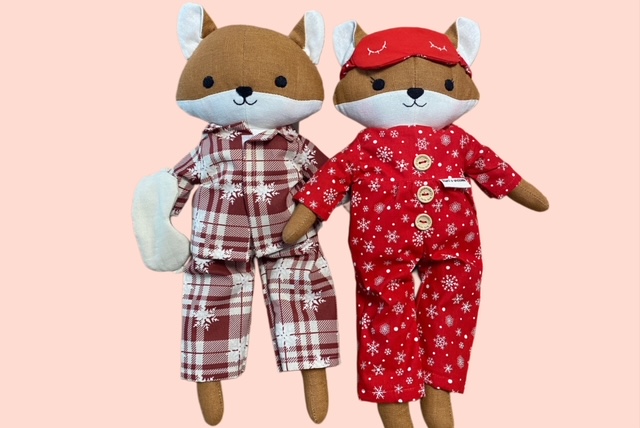 Zwei Stofffüchse in Winter-Pyjamas einer im rot-beigen Karomuster, der andere im roten Schneeflocken-Strampler mit Schlafmaske aus rosa Hintergrund.