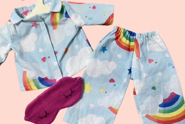 Nahaufnahme von Handgenähten Puppen-Pyjama-Set mit Regenbogen und Wolkenmuster im Blau kombiniert mit einer pinken Schlafmaske.