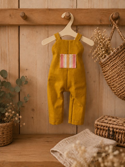 Handgenähte gelbe Puppen- Latzhose mit kleiner gestreifter Tasche an einer rustikalen Holzwand dekoriert.