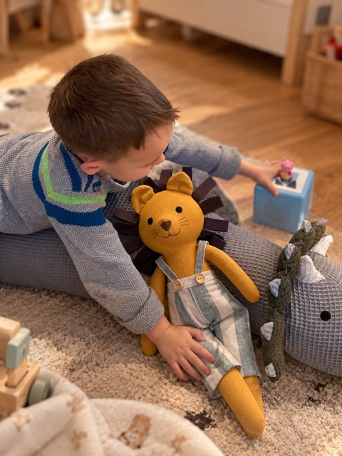 Kleiner Junge spielt auf dem Kinderzimmerteppich mit einem handgefertigten Löwen- Begleiter aus Leinen mit brauner Schlaufenmähne. 