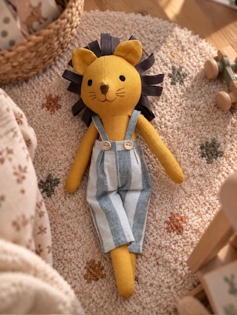 Handgefertigter Leinenlöwen mit braunen Stoffschlaufen zum Fühlen und Greifen, liegend auf einem weichen Spielteppich im Kinderzimmer.